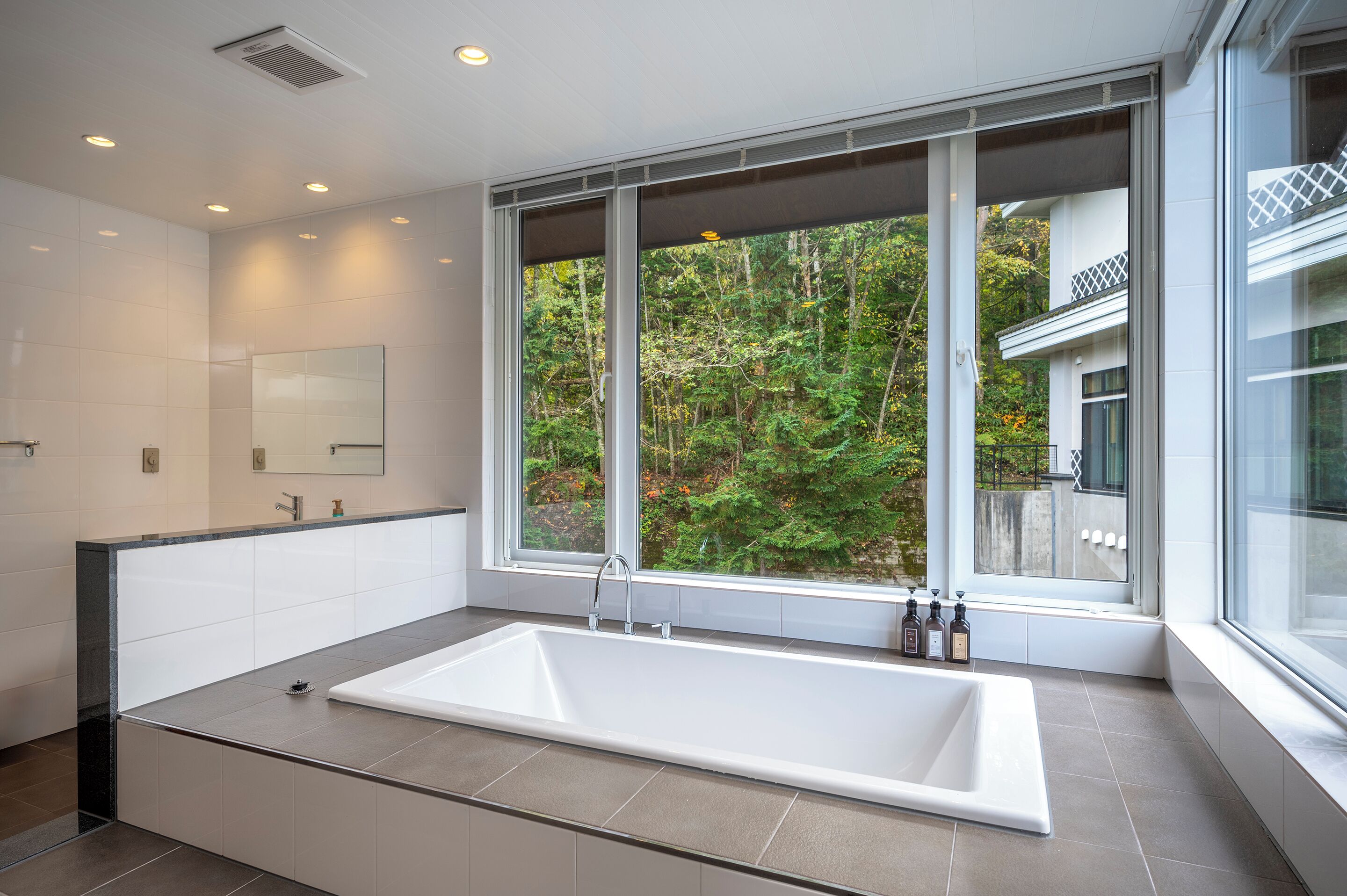 A modern bathroom with a large bathtub, a mirror, and a window offering a view of a forest.