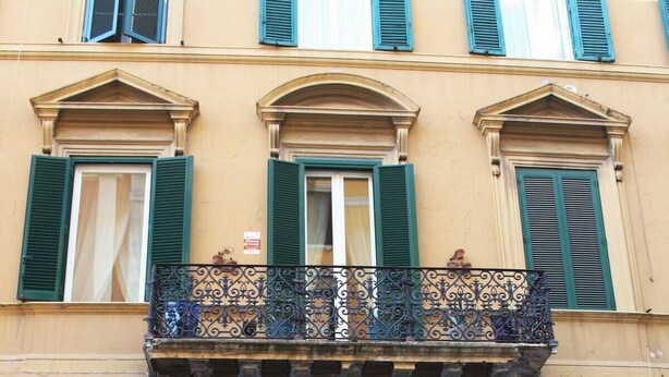 Um edifício com entrada central, venezianas verdes e uma varanda com grades de metal decoradas.