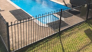 A rectangular swimming pool surrounded by a black metal fence, with a covered seating area and a grassy area in the foreground.