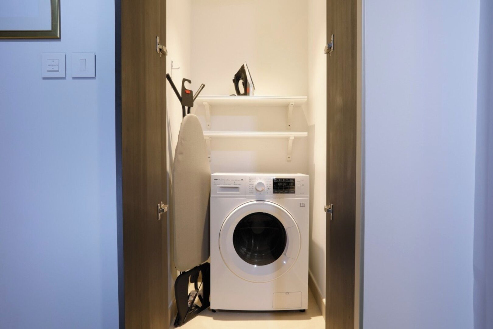 A compact laundry room with a front-loading washing machine, a shelf above it, and a door leading to another room.