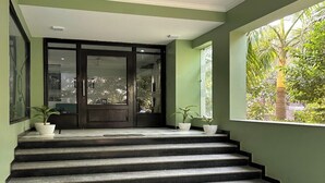 Een trap die leidt naar een donker houten deur met glaspanelen, omlijst door potplanten en een groot raam met uitzicht op groen.