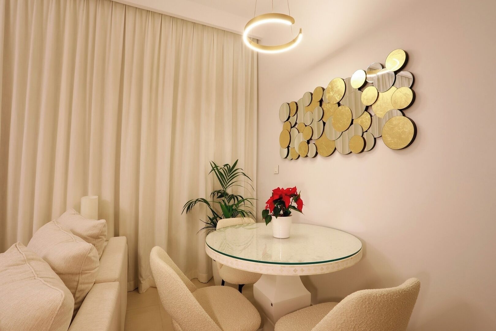 A modern living room with a round glass table, beige sofas, a potted plant, and a decorative wall piece.