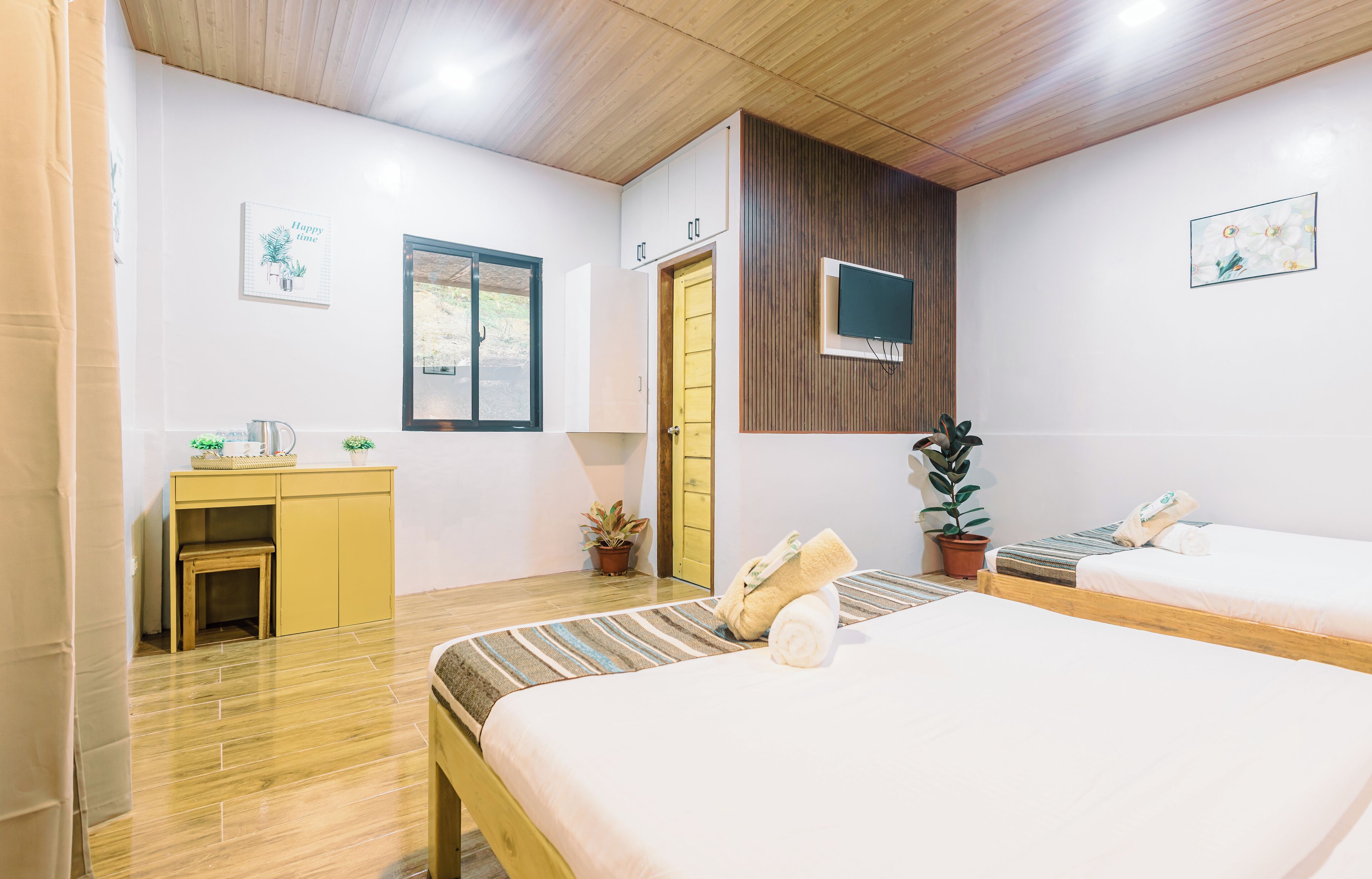 Une chambre d’hôtel avec deux lits, un plafond en bois, un téléviseur fixé au mur et un petit bureau agrémenté d’une plante.