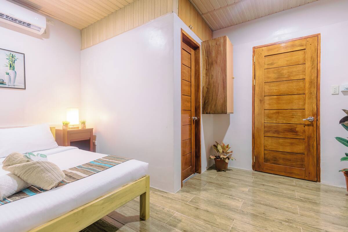 A hotel room with a bed, bedside table, lamp, wooden wardrobe, and a door.