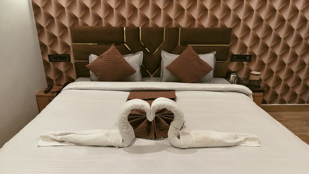 A neatly made bed with a heart-shaped towel arrangement, a brown headboard, and a patterned wall. - The Kashi Yatra Inn (Varanasi)