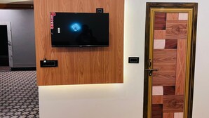 A modern room with a mounted flat-screen TV on a wooden panel, a door with a geometric wood pattern, and a patterned floor. - The Kashi Yatra Inn (Varanasi)