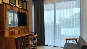 Een moderne woonkamer met een flatscreen televisie, houten meubels en grote ramen met dunne gordijnen.