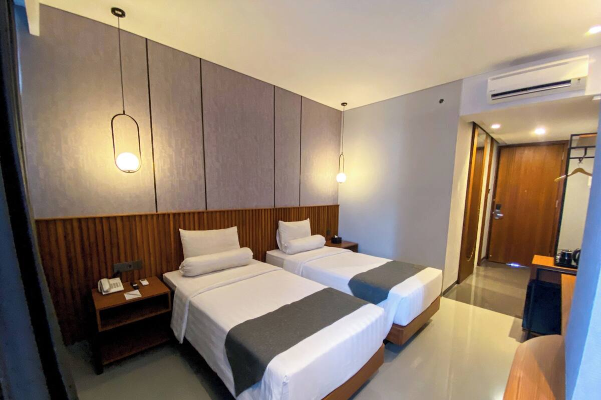 A hotel room with two beds, wooden headboards, bedside tables, and a desk with a chair.