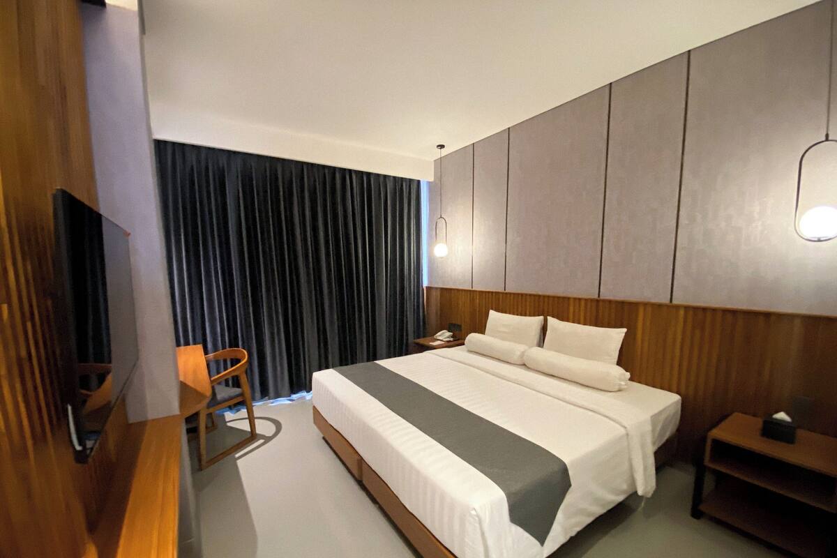 A modern hotel room with a large bed, a wooden desk, a chair, a nightstand, and a large window with curtains.