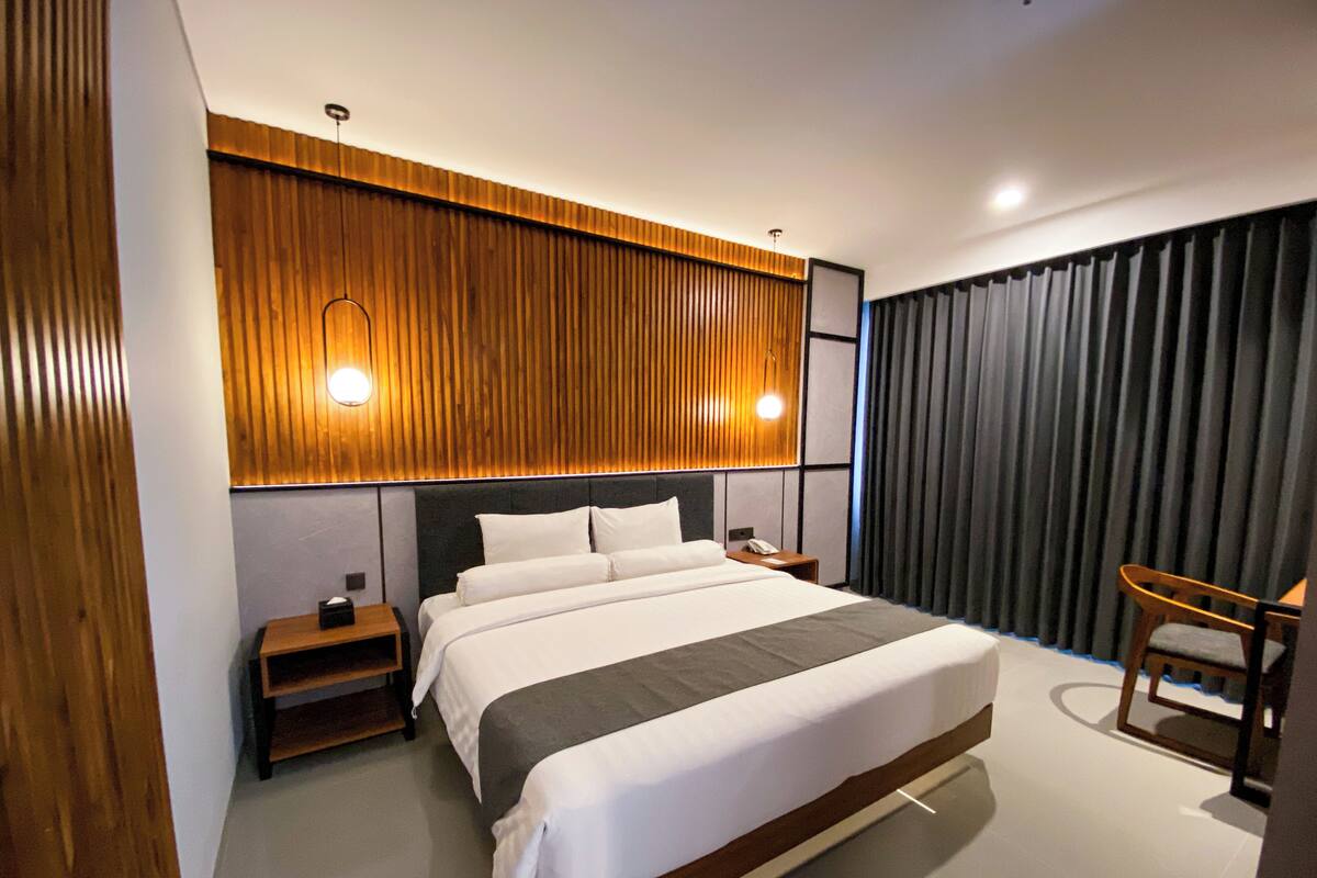 A modern hotel room with a large bed, wooden panel wall, two bedside tables, a chair, and a large window with curtains.