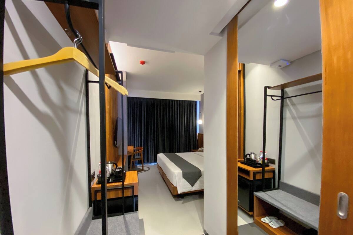 A modern hotel room with a bed, a desk with a chair, a small table with a kettle, and a mirror.