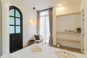 Ett hotellrum med en säng, en stol, en hylla med diverse föremål och ett fönster med gardiner.