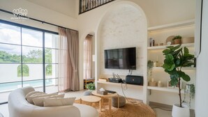 A modern living room with a large TV, comfortable seating, a fireplace, and a view of greenery. - Oxygen Valley Khao Yai B2 (Pak Chong)