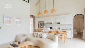 A modern living room with a sofa, coffee table, and chairs. There is a kitchen area with white cabinets and a dining table. - Oxygen Valley Khao Yai B2 (Pak Chong)