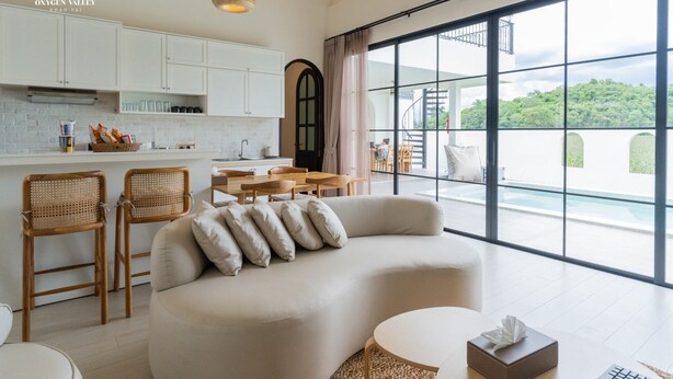A modern living room with a sofa, two wicker chairs, a dining area, and large windows offering a view of a pool and greenery. - Oxygen Valley Khao Yai B2 (Pak Chong)