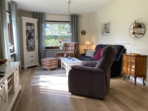 Una sala de estar con sofá, sillón, banqueta, aparador de madera y una ventana con cortinas.