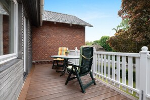 A wooden deck with a table and chairs, a white fence, and a brick building in the background.