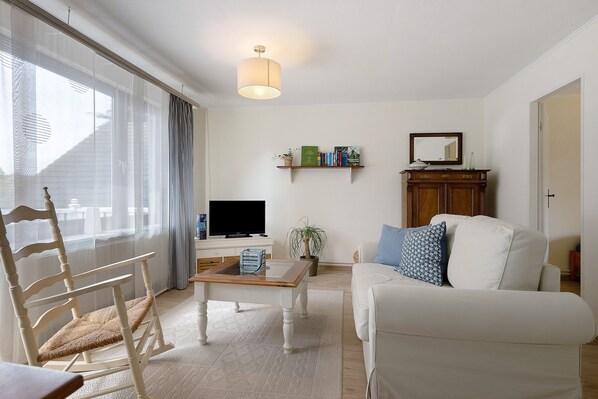 A living room with a white sofa, a wooden chair, a coffee table, a TV, a bookshelf, and a potted plant.