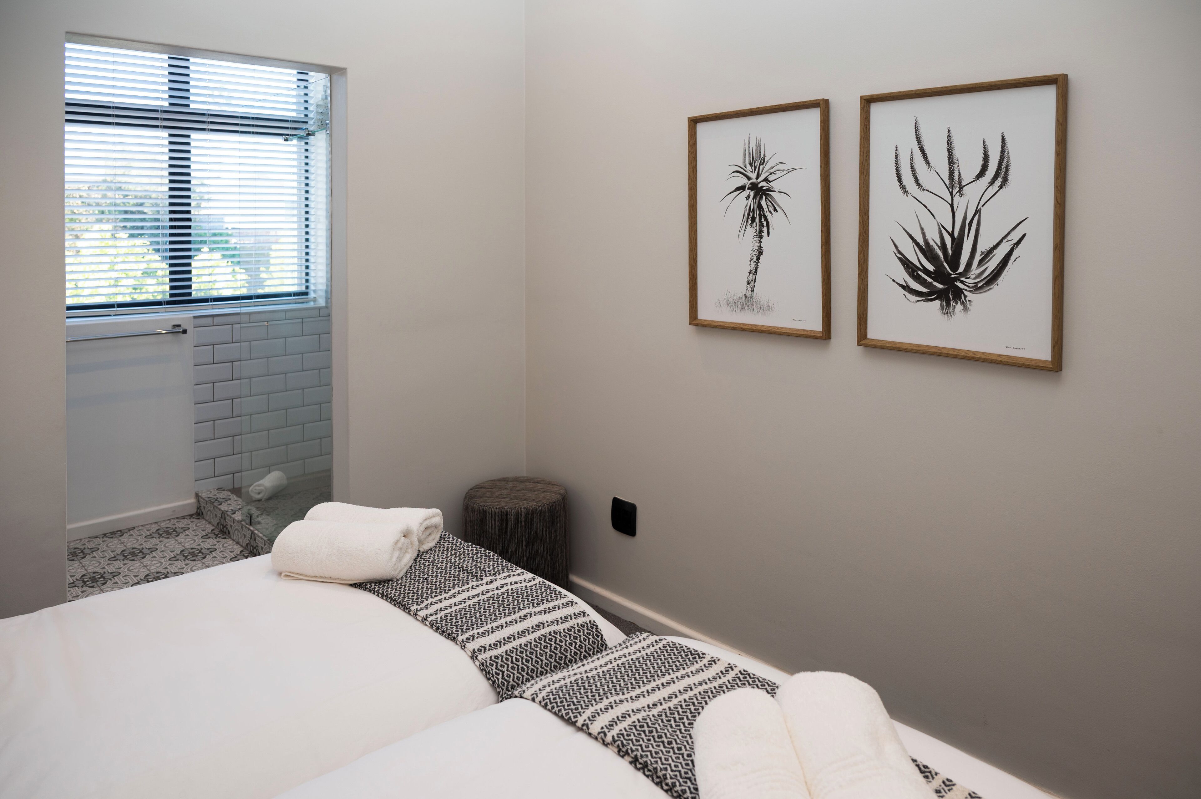 Ein Schlafzimmer mit einem Bett, einem Fenster mit Jalousien und zwei gerahmten Pflanzenbildern an der Wand.