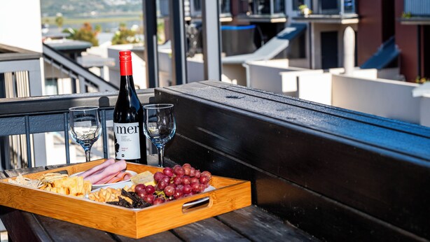 Una bandeja de madera con aperitivos y una botella de vino Mer en un balcón con vistas a edificios y colinas.