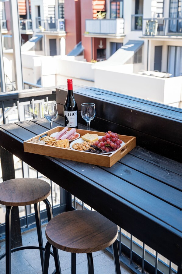 A wooden table with a tray of snacks, two glasses of wine, and a bottle of wine on a balcony.