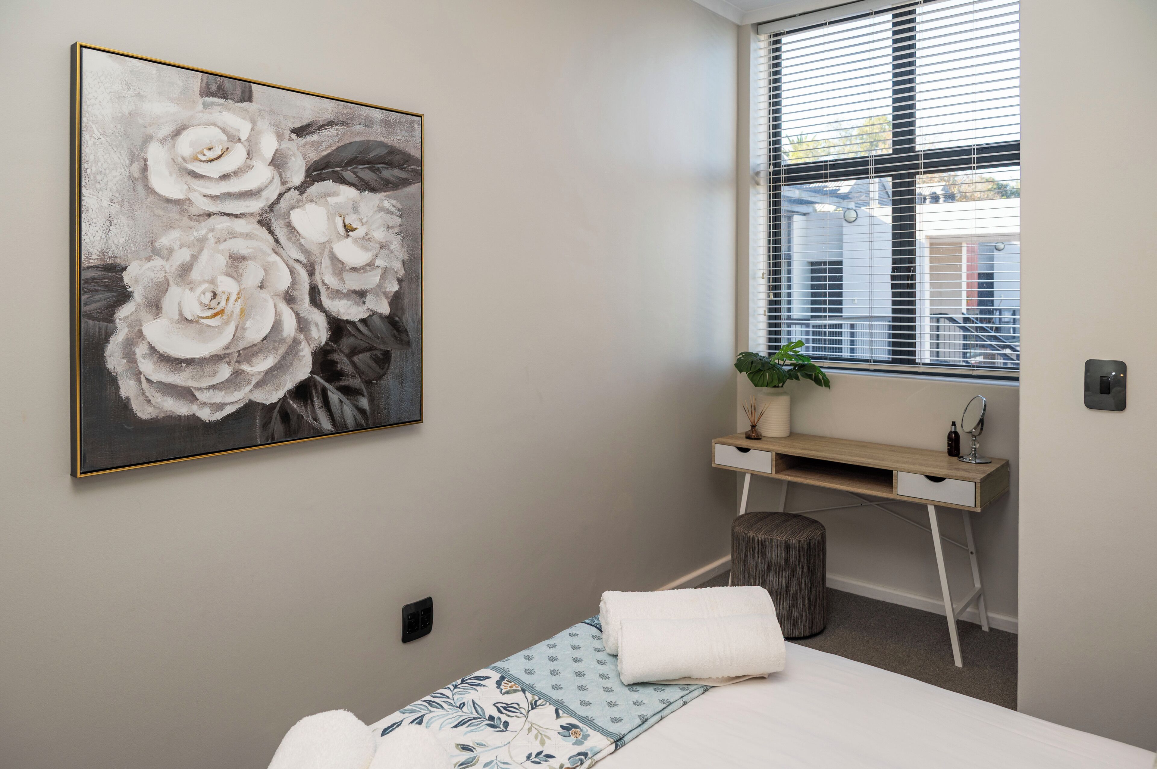 A bedroom with a bed, a desk, a potted plant, and a large floral painting on the wall.