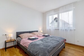 Une chambre à coucher avec un lit, des tables de chevet, une fenêtre avec des rideaux légers et une vue sur une maison à l’extérieur.