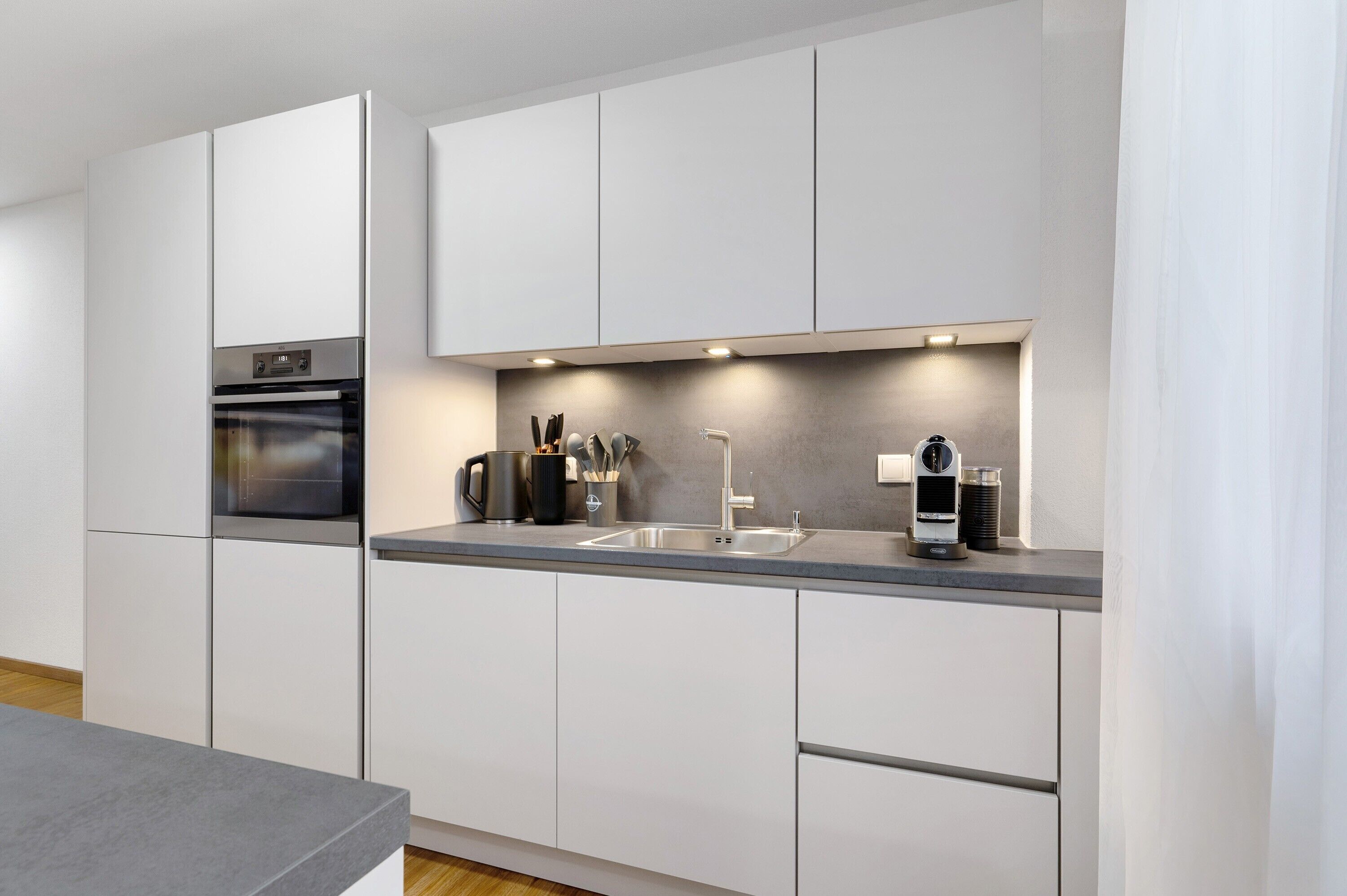 A modern kitchen with white cabinetry, a built-in oven, a stainless steel sink, and under-cabinet lighting.