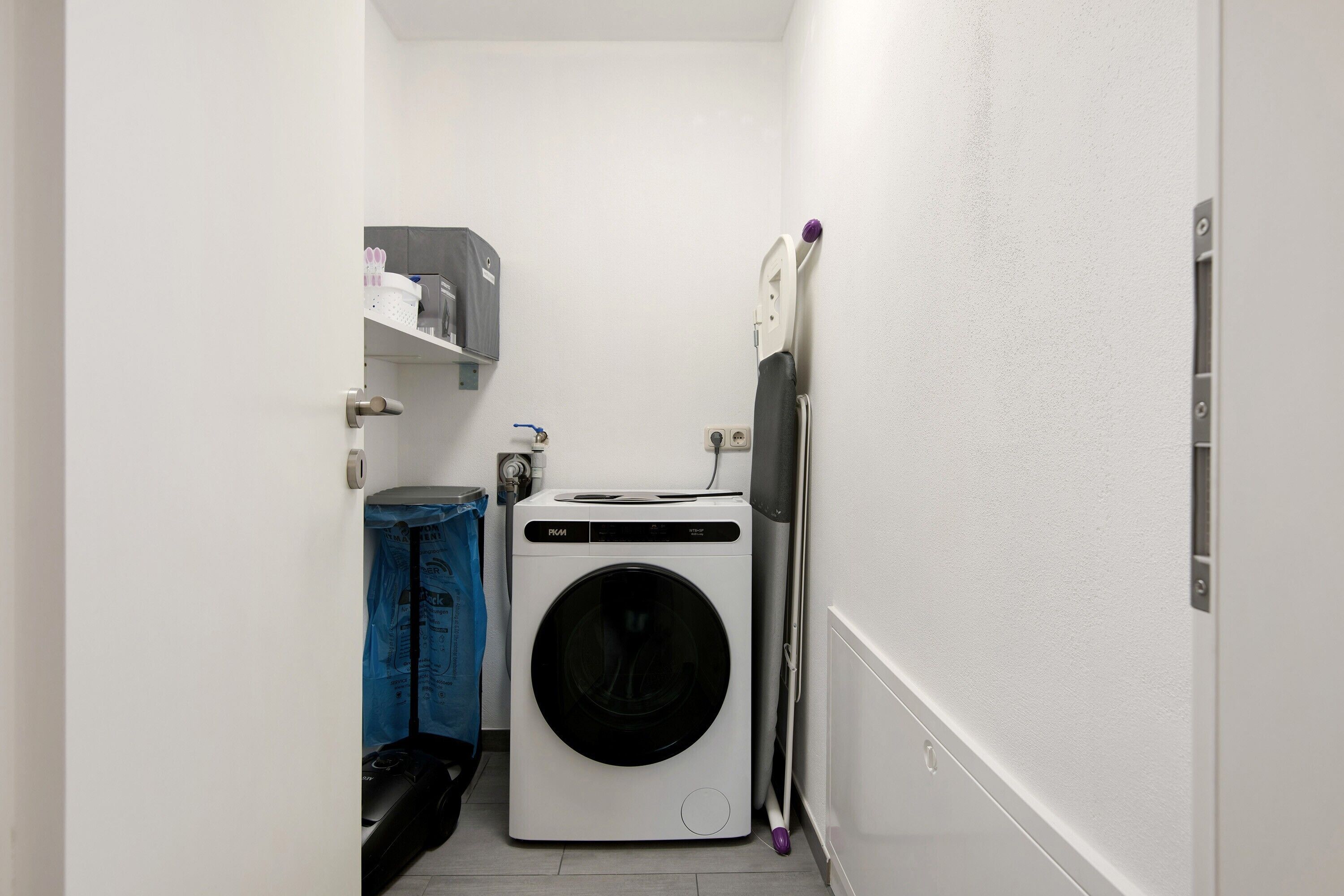 Une petite buanderie équipée d’un lave-linge à chargement frontal, d’un étendoir et d’une étagère contenant le nécessaire pour faire la lessive.