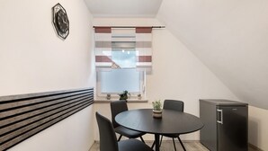 Una habitación compacta con una mesa redonda, cuatro sillas, un refrigerador pequeño y una ventana con persianas.
