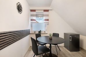 Quarto compacto com uma mesa redonda, quatro cadeiras, um frigorífico pequeno e uma janela com persianas.