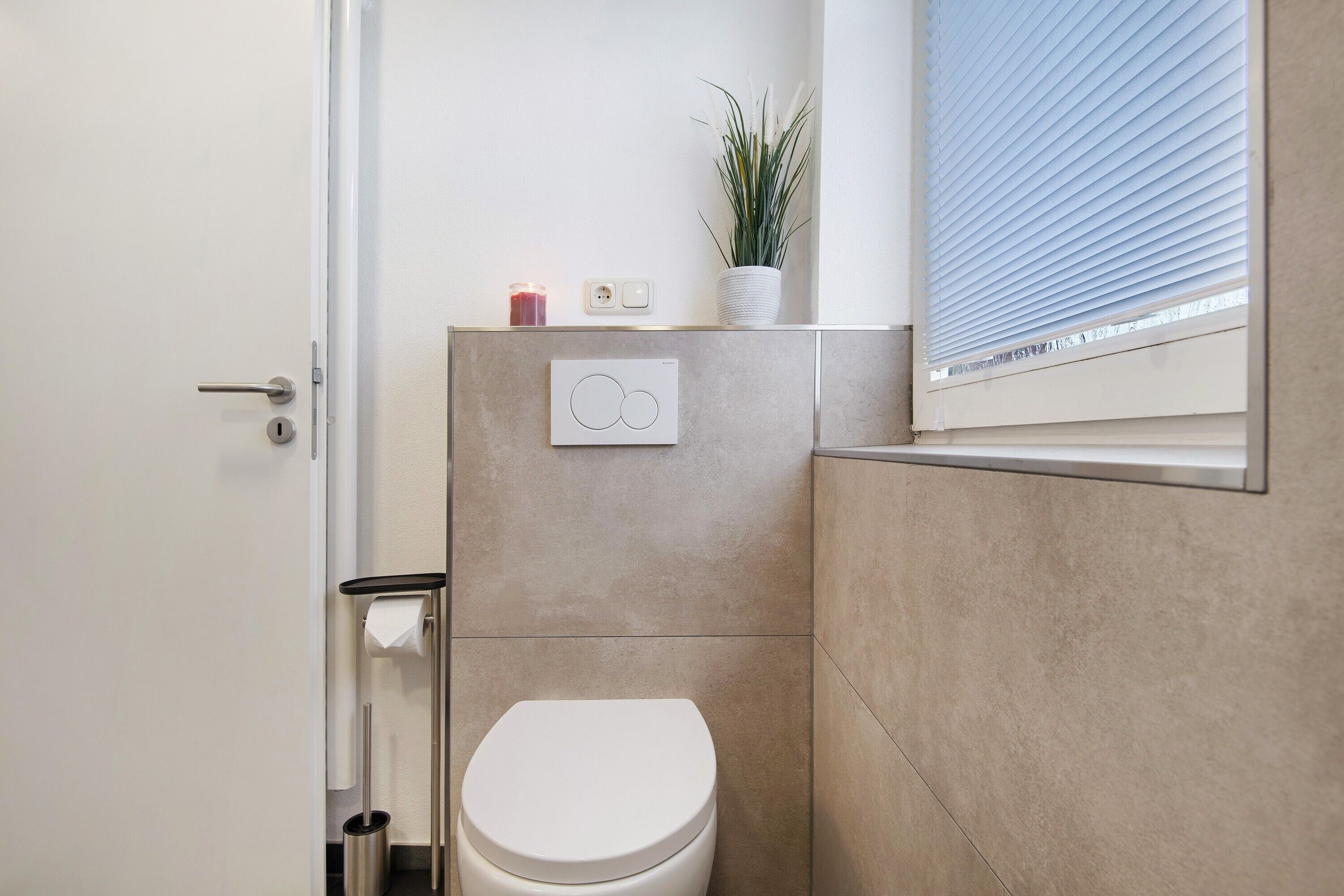 Moderni kylpyhuone, jossa on wc, käsienkuivaaja, roskakori, seinään asennettu valokytkin ja ikkuna, jossa on sälekaihtimet.