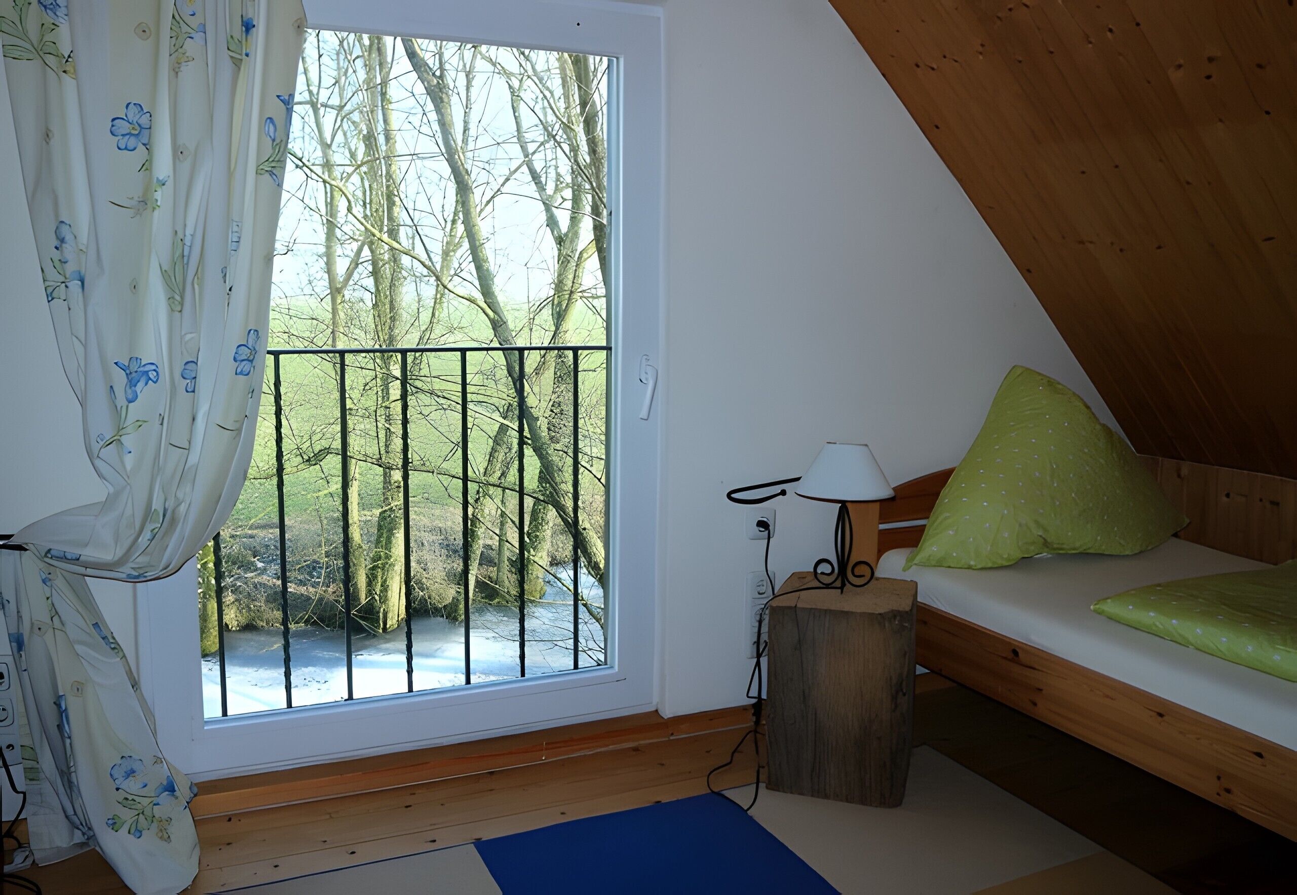 Ein Schlafzimmer mit einem hölzernen Bett, einem Fenster mit Blick auf Bäume und einen Fluss, sowie einem Vorhang mit einem Blumenmuster.
