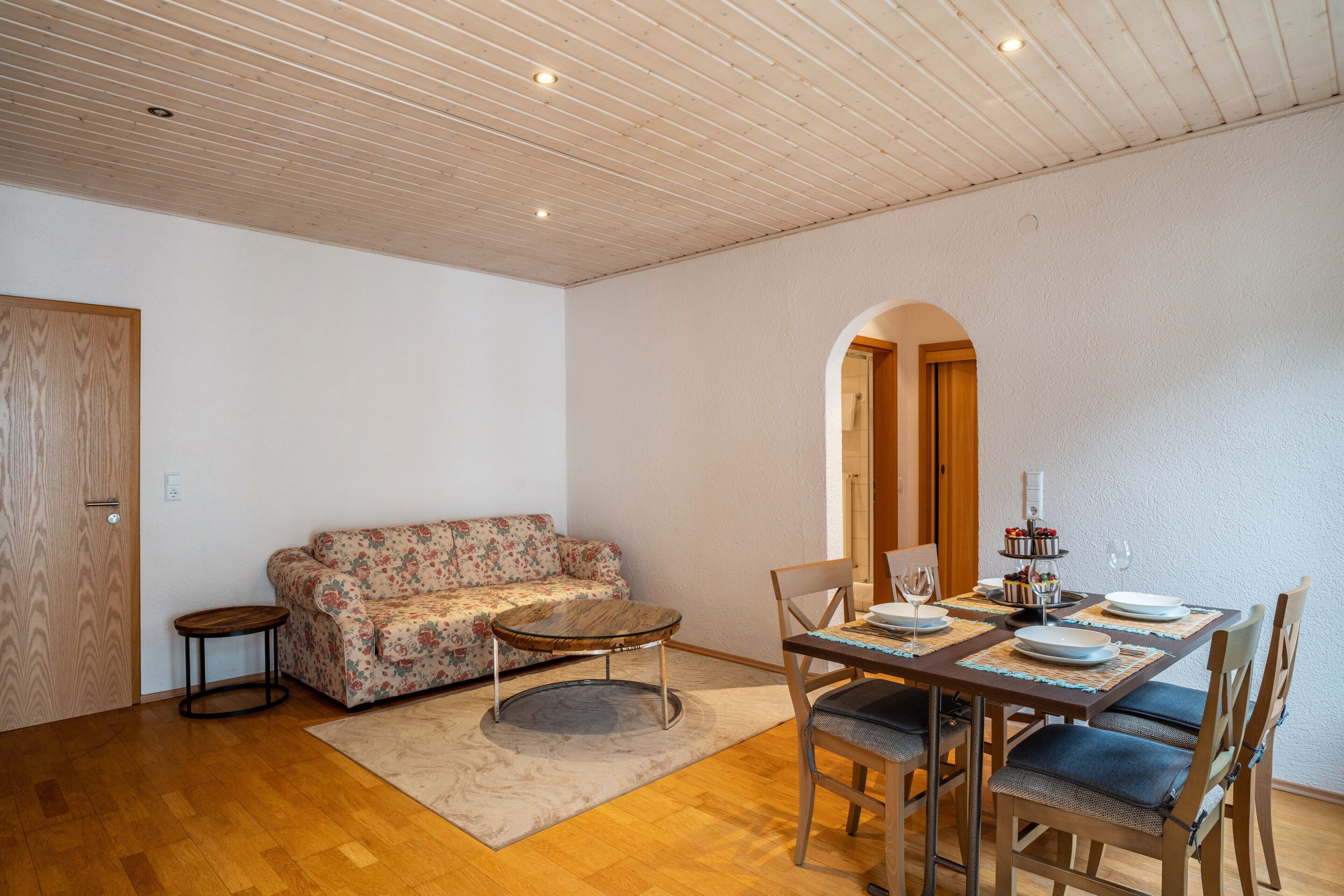 Un comedor con una mesa de madera dispuesta para cuatro personas, un sofá floral y una pequeña mesa redonda.