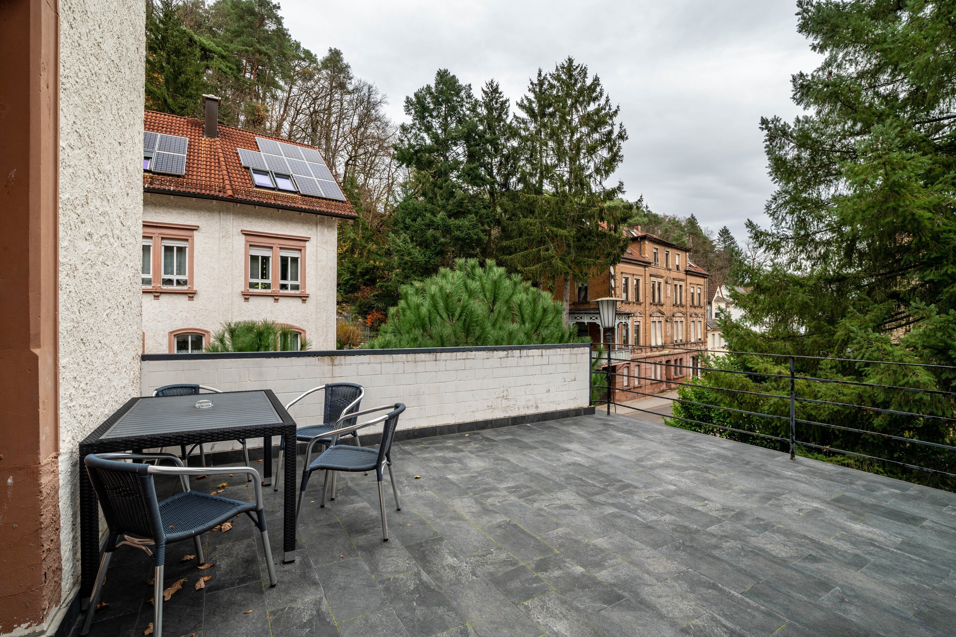 Eine Terrasse mit Tisch und Stühlen, eine weiße Wand und Blick auf Gebäude und Bäume.