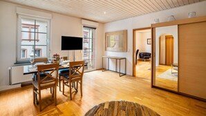 A modern living room with a wooden floor, a dining table with chairs, a flat-screen TV, and a large window with a view of buildings.
