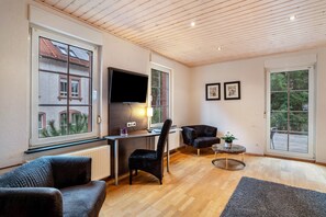 A modern living room with a wooden ceiling, a desk with a computer, a black chair, a sofa, and large windows.