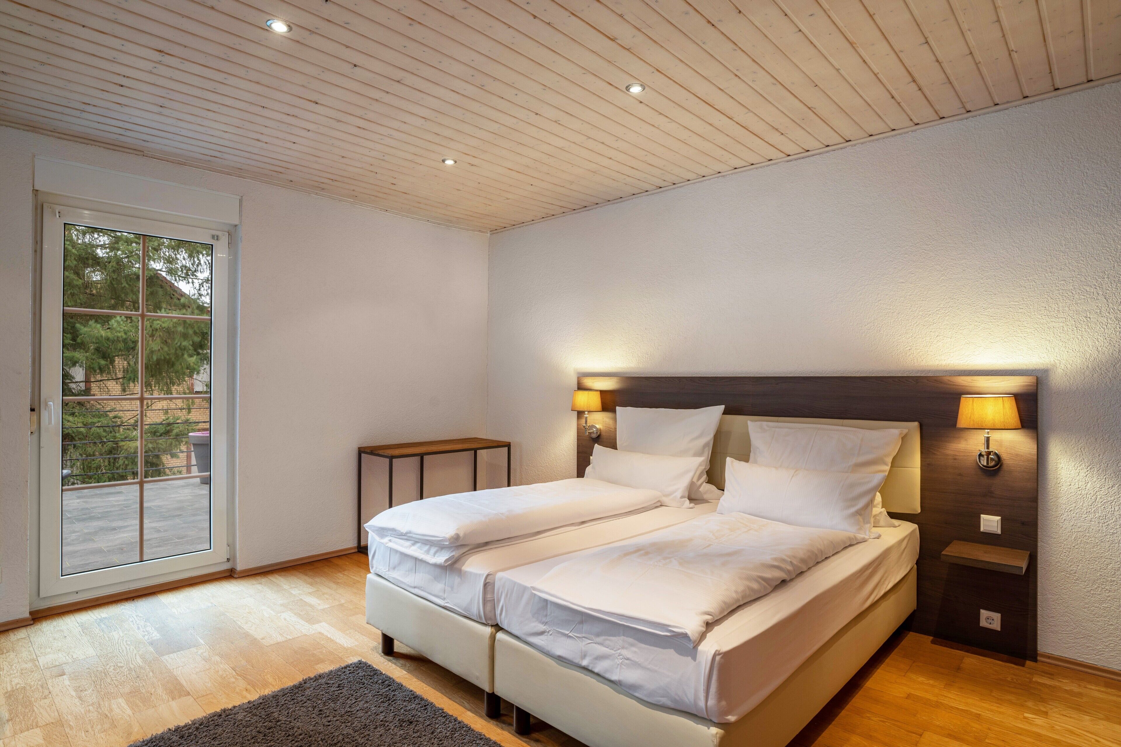 Quarto com uma cama de casal, duas mesas de cabeceira, um criado-mudo, uma porta de vidro deslizante e teto de madeira.