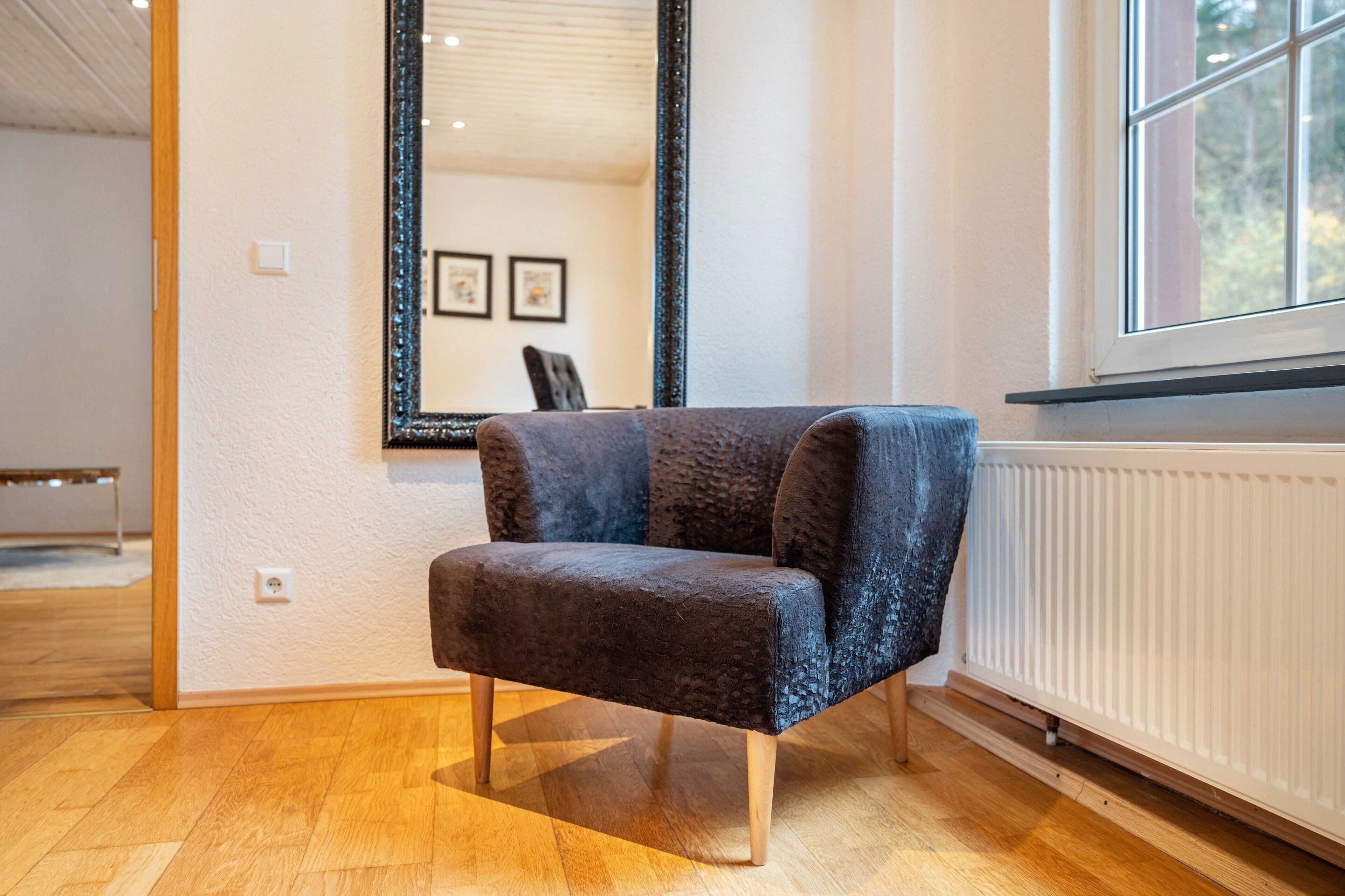 Une pièce avec un plancher en bois, un fauteuil à l’assise sombre, un grand miroir et une fenêtre donnant sur des arbres.