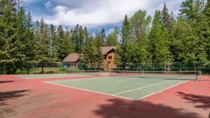 Una cancha de tenis al aire libre con una red, rodeada de árboles y una cabaña al fondo.
