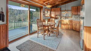 A spacious kitchen with wooden cabinets and a large island, a dining area with a table and chairs, and a sliding glass door leading to a balcony with a view of trees. - The Eagles Nest – Couples Escape - Hot Tub & Views! (Ellicottville)