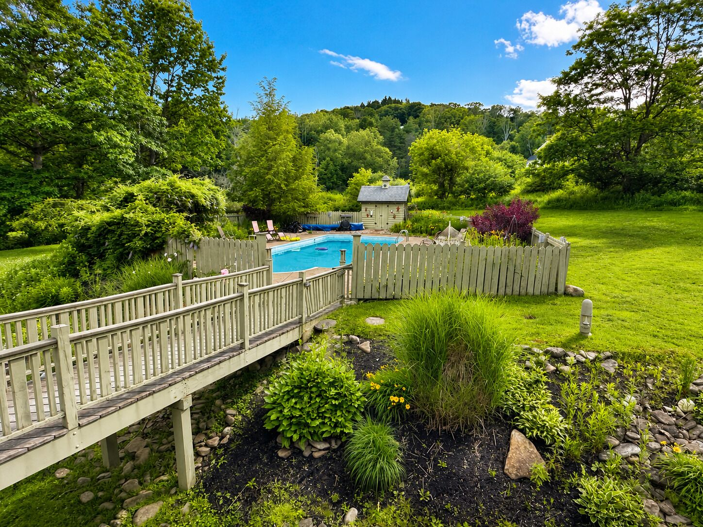A backyard with a swimming pool, wooden deck, and lush greenery.