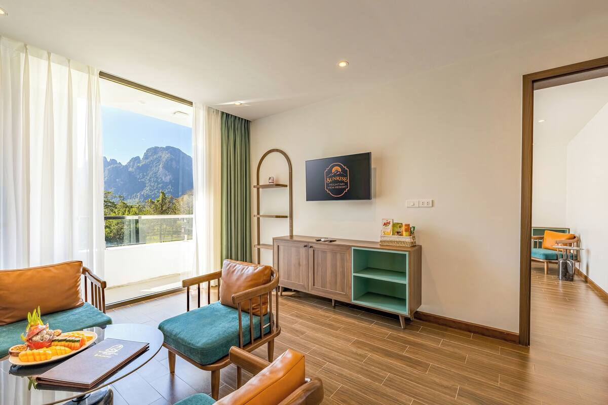 A modern hotel room with a large window offering a mountain view, a flat-screen TV mounted on the wall, and a wooden cabinet with a green shelf.