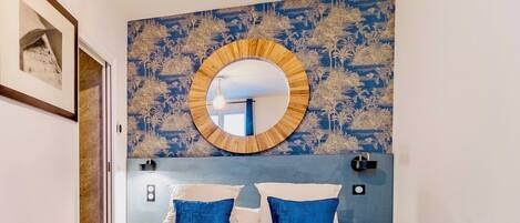 A bedroom with a bed, two blue pillows, a round mirror, and a wall with a floral pattern.