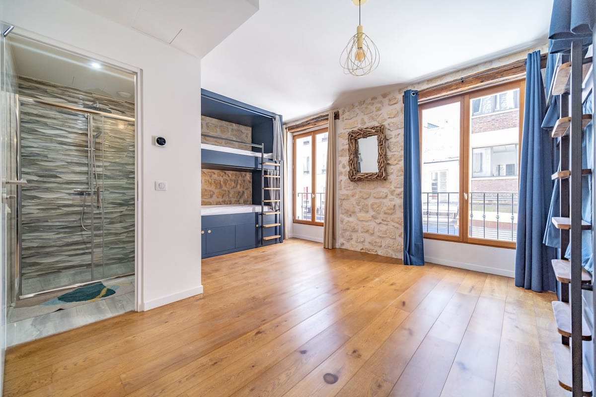Une pièce spacieuse dotée d’un plancher en bois, d’une cabine de douche en verre, d’un banc intégré avec espace de rangement et d’une grande fenêtre donnant sur des immeubles.