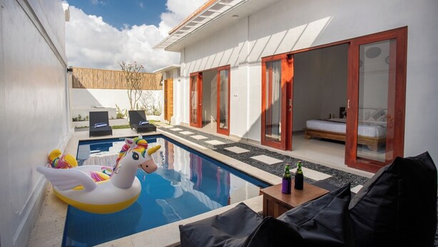 Zona de piscina exterior moderna con un unicornio inflable blanco, sillones de descanso y una mesa con botellas.
