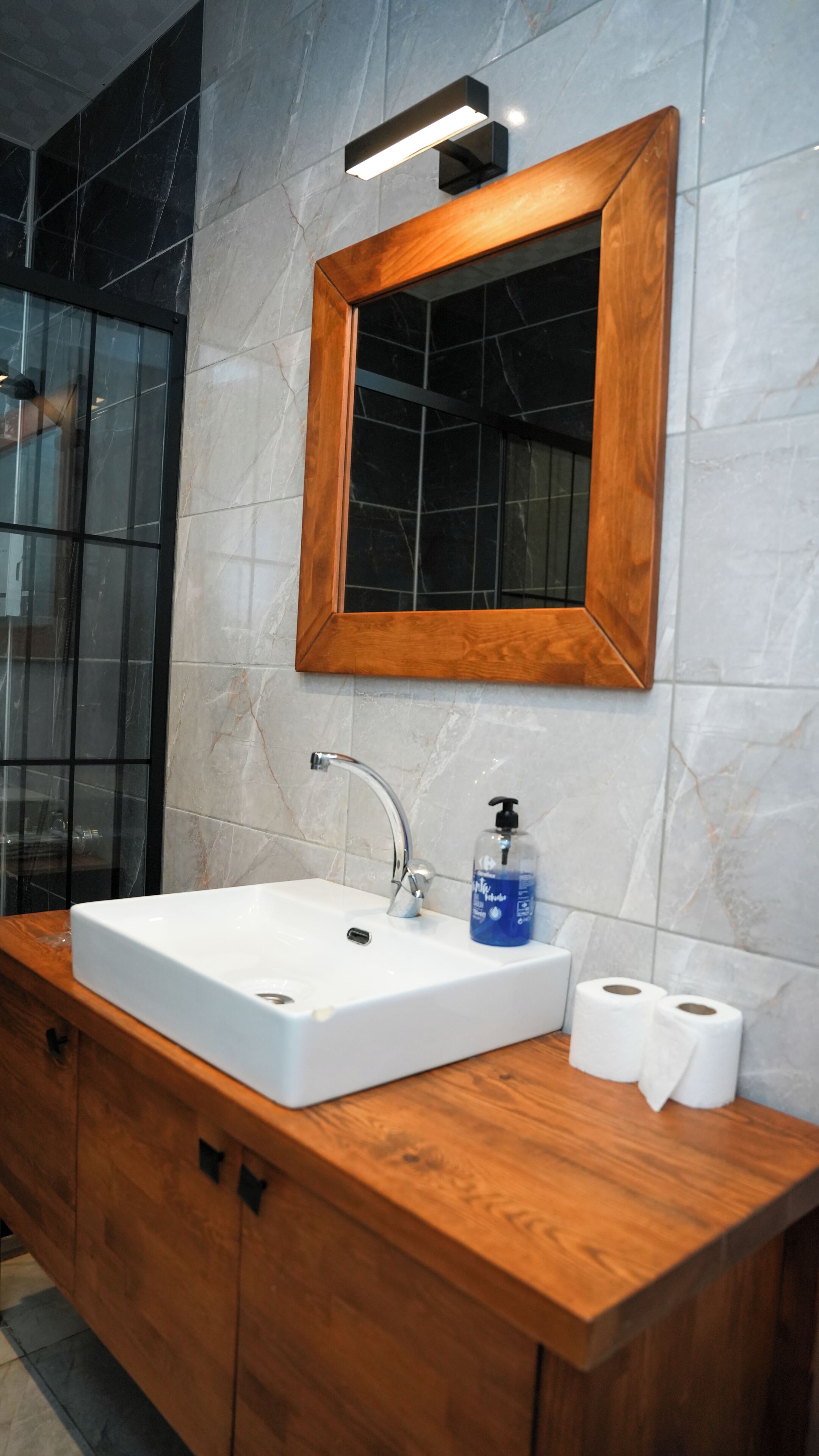 A bathroom with a wooden vanity, a white sink, a mirror, and two rolls of toilet paper.