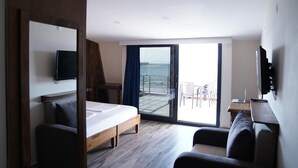 A hotel room with a bed, a desk with a laptop, a TV mounted on the wall, and a balcony with a view of the sea. - ASTROİA OTEL & BİLİM KAMPI (Lapseki)