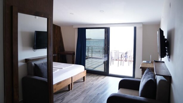 A hotel room with a bed, a desk with a laptop, a TV mounted on the wall, and a balcony with a view of the sea. - ASTROİA OTEL & BİLİM KAMPI (Lapseki)