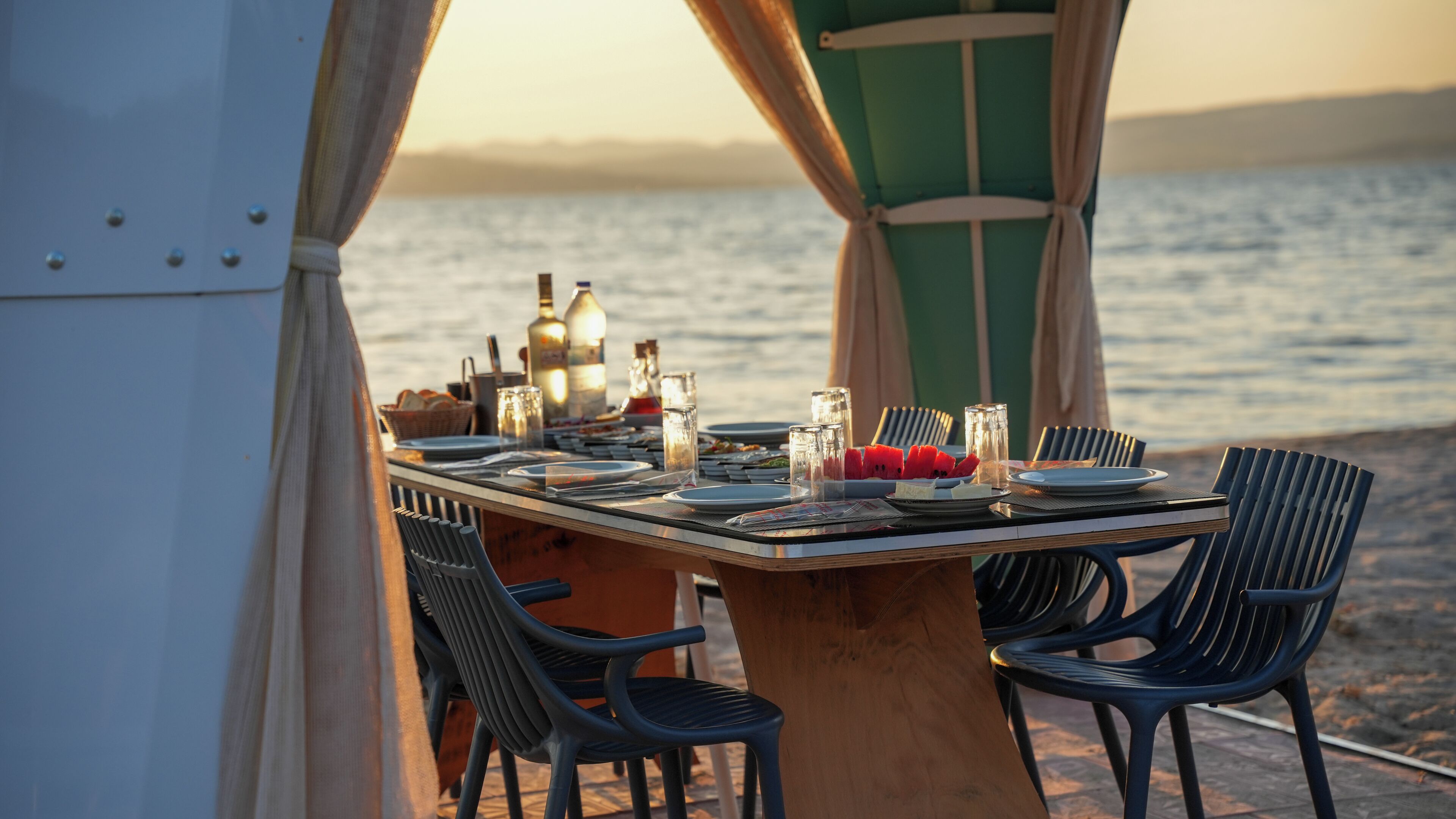Mesa de comedor al aire libre con sillas, platos, cubiertos y vasos, con vista a un cuerpo de agua.
